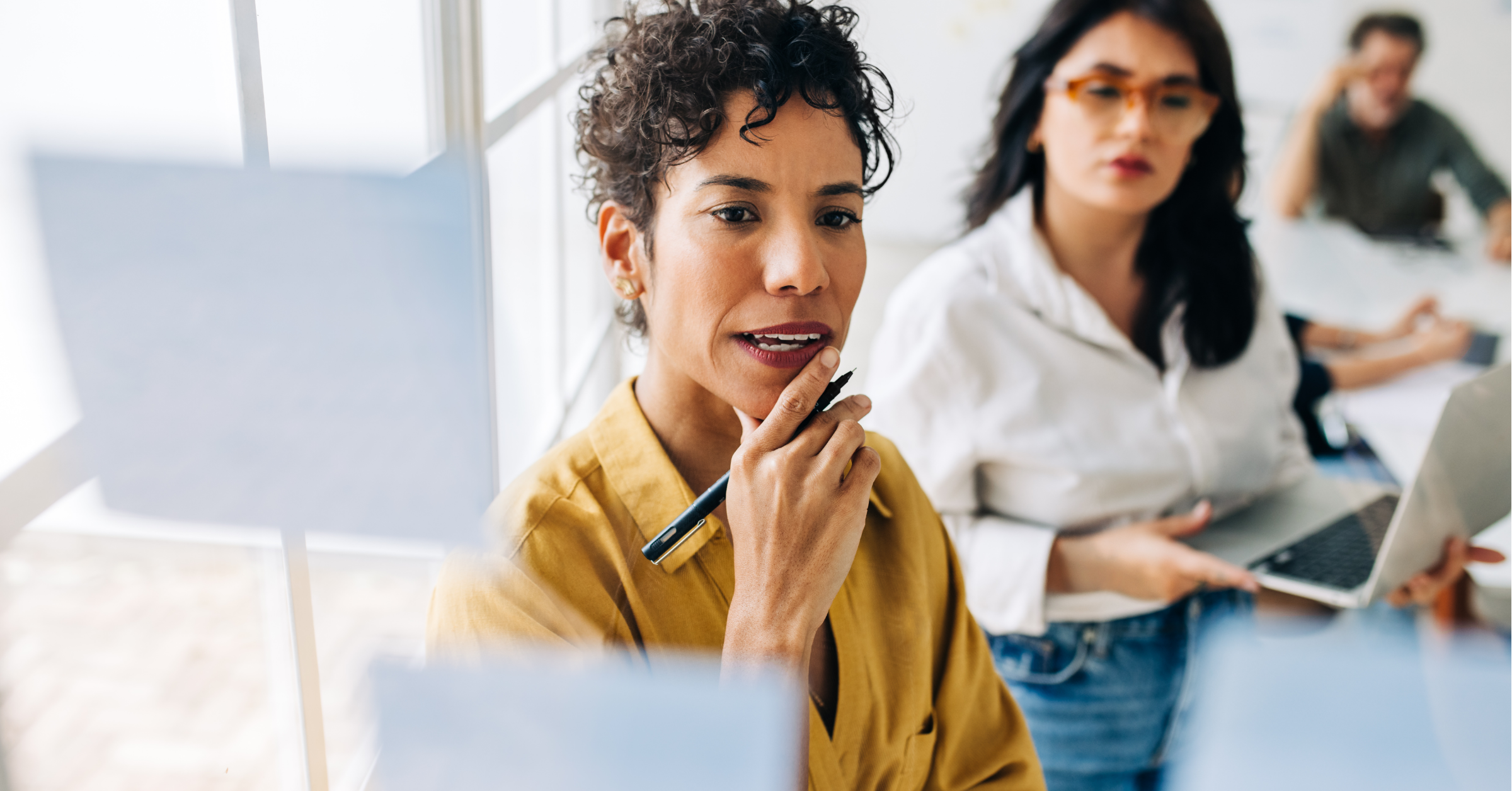 woman with a yellow shirt thinking with her hand on her chin holding a pen in a business setting.