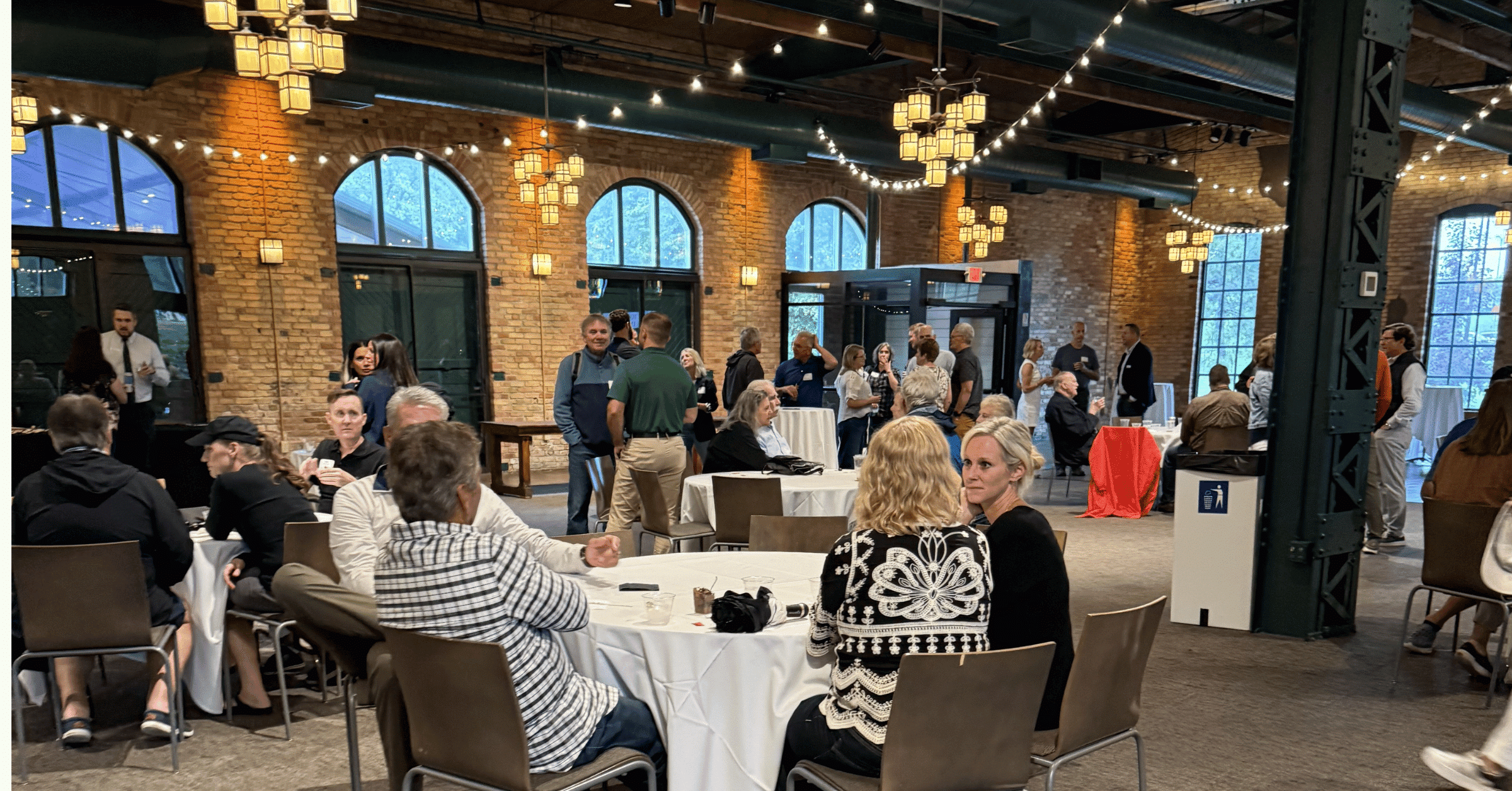 People sitting around a table enjoying conversation in an event space with cafe lights, large window and brick walls.